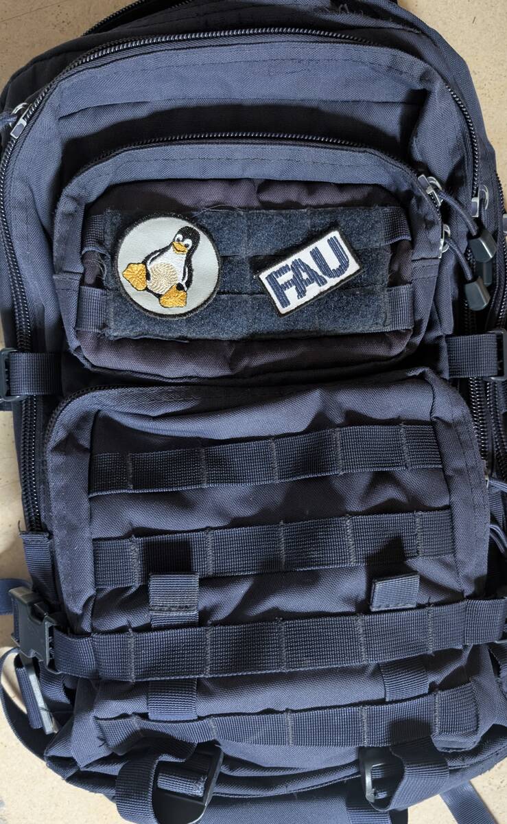 Dunkelblauer Rucksack im Military-Stil, auf dessen schwarzer Flauschfläche ein runder Tux-Patch und ein rechteckiger FAU-Patch haften